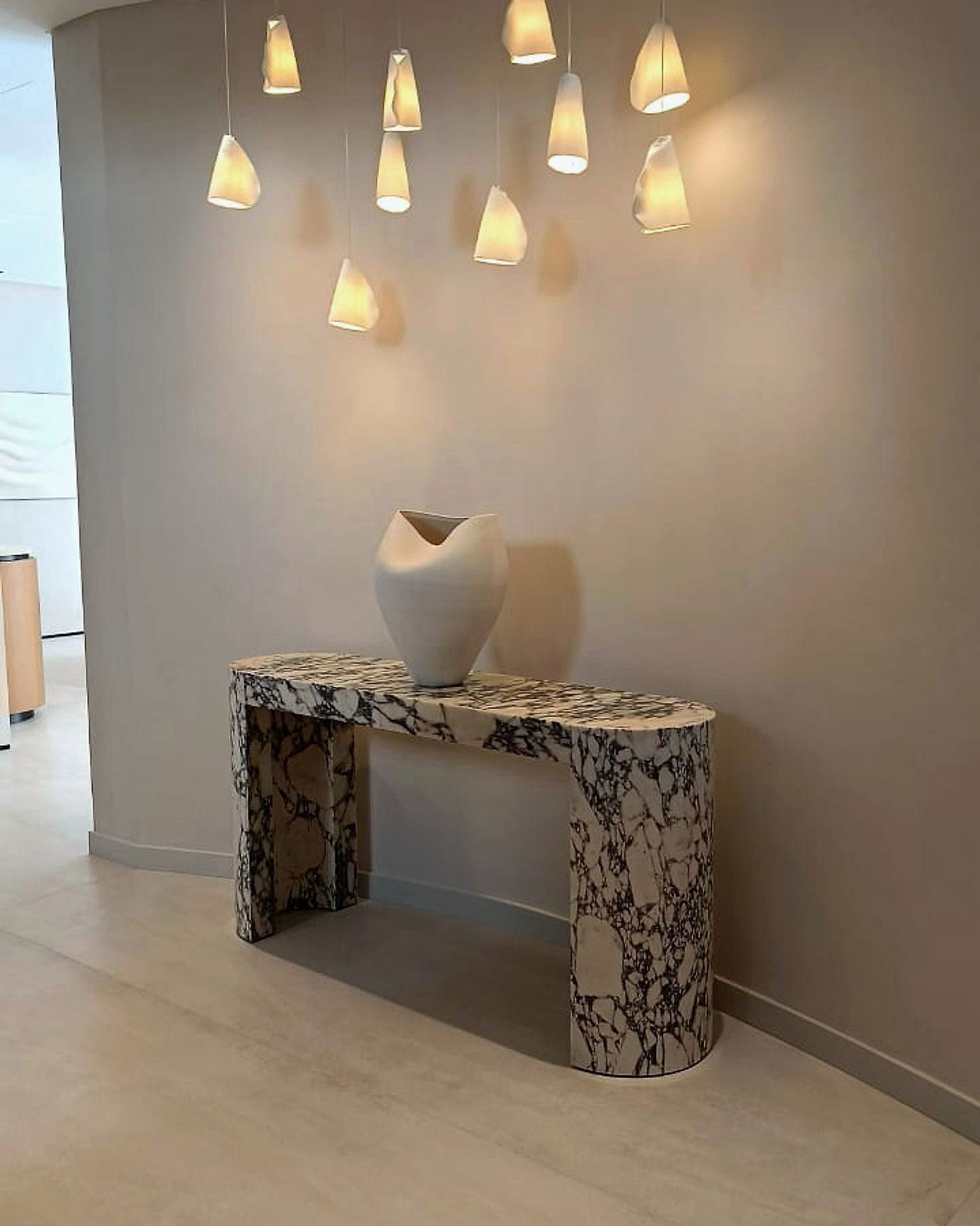 A Viola marble console on a curved micro-cement wall, lit by a scattered cluster of alabaster pendants.