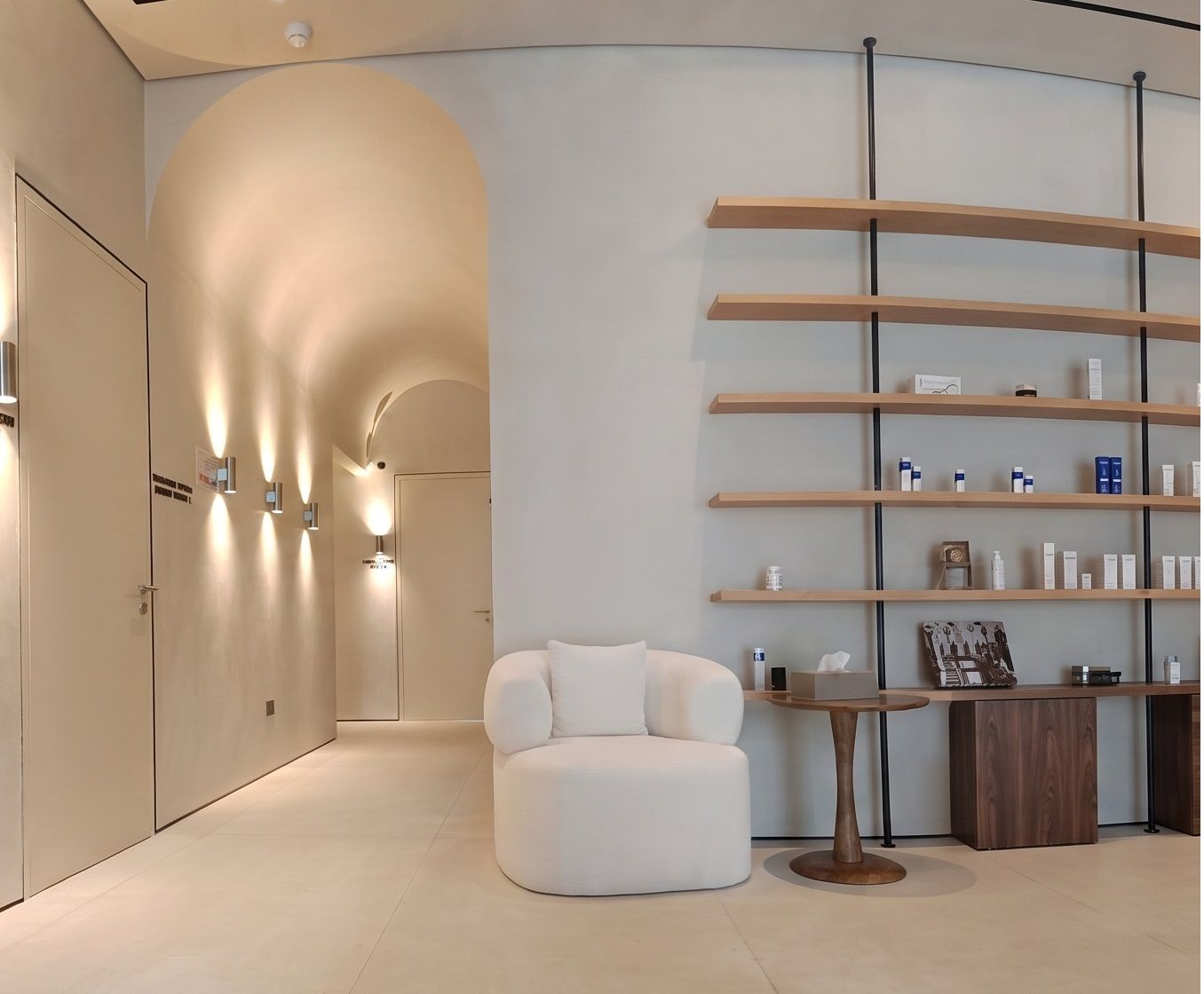 Oak shelves on blackened steel uprights beside a barrel-vaulted corridor washed by wall lights, with a bouclé chair between the two.