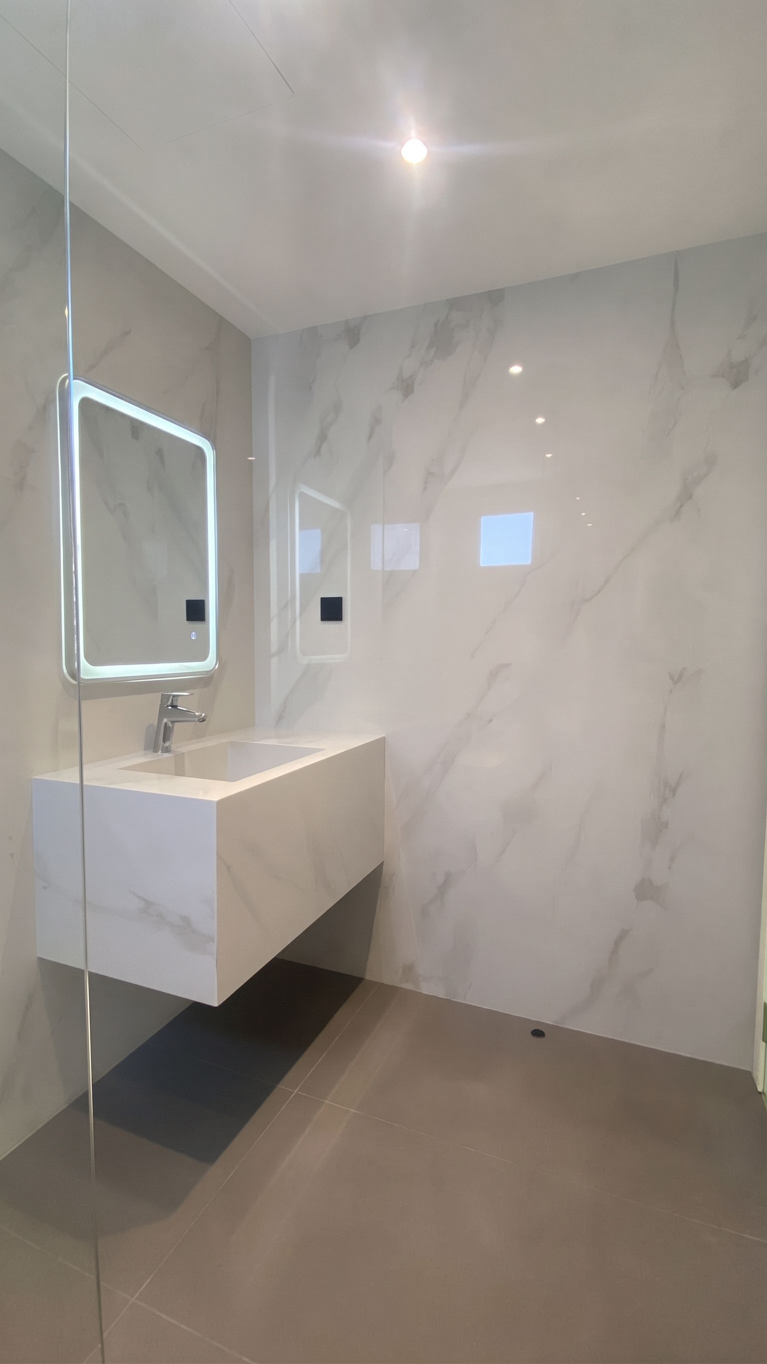 A cantilevered vanity with a moulded basin and backlit mirror, with a frameless glass fin dividing off the wet area.