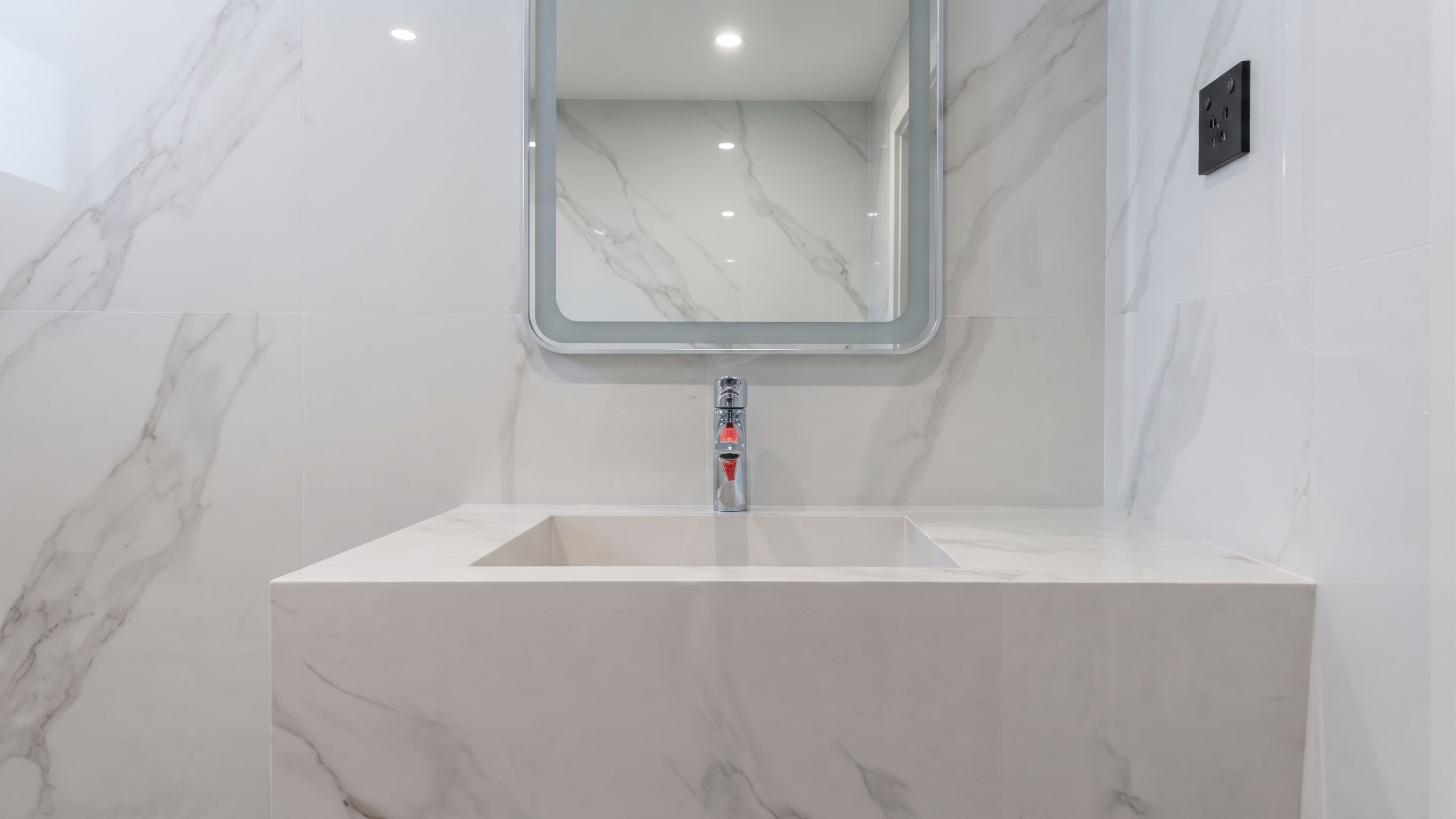 A solid basin block with the bowl cut into the top, set against book-matched marble-effect panels under a backlit mirror.