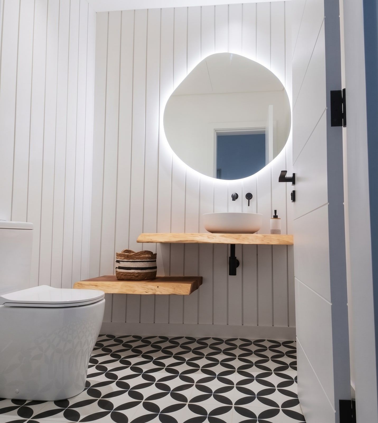 An organic backlit mirror on fluted panelling, with a floating timber shelf, vessel basin and patterned cement floor.