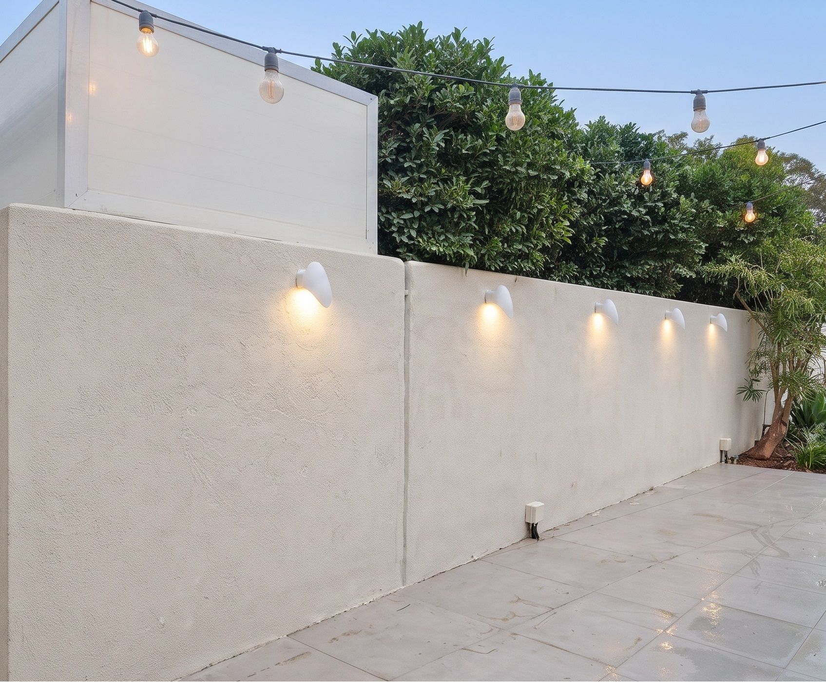 Sculptural uplighters set along a rendered garden wall, with festoon lighting over the paving.