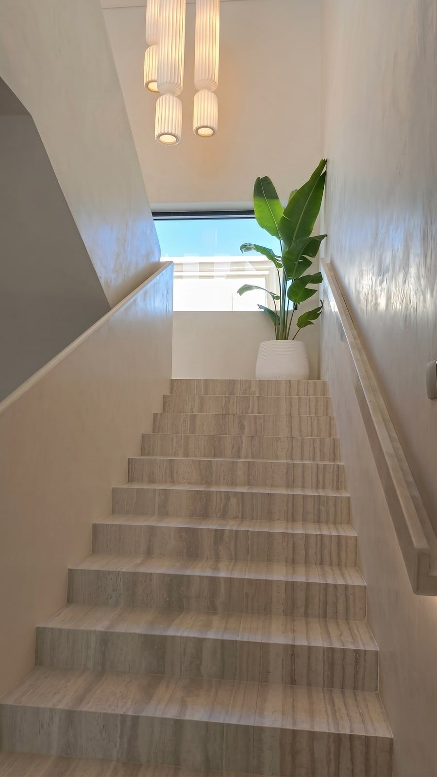 Vein-cut travertine treads with a solid stone handrail and a fluted pendant cluster overhead.